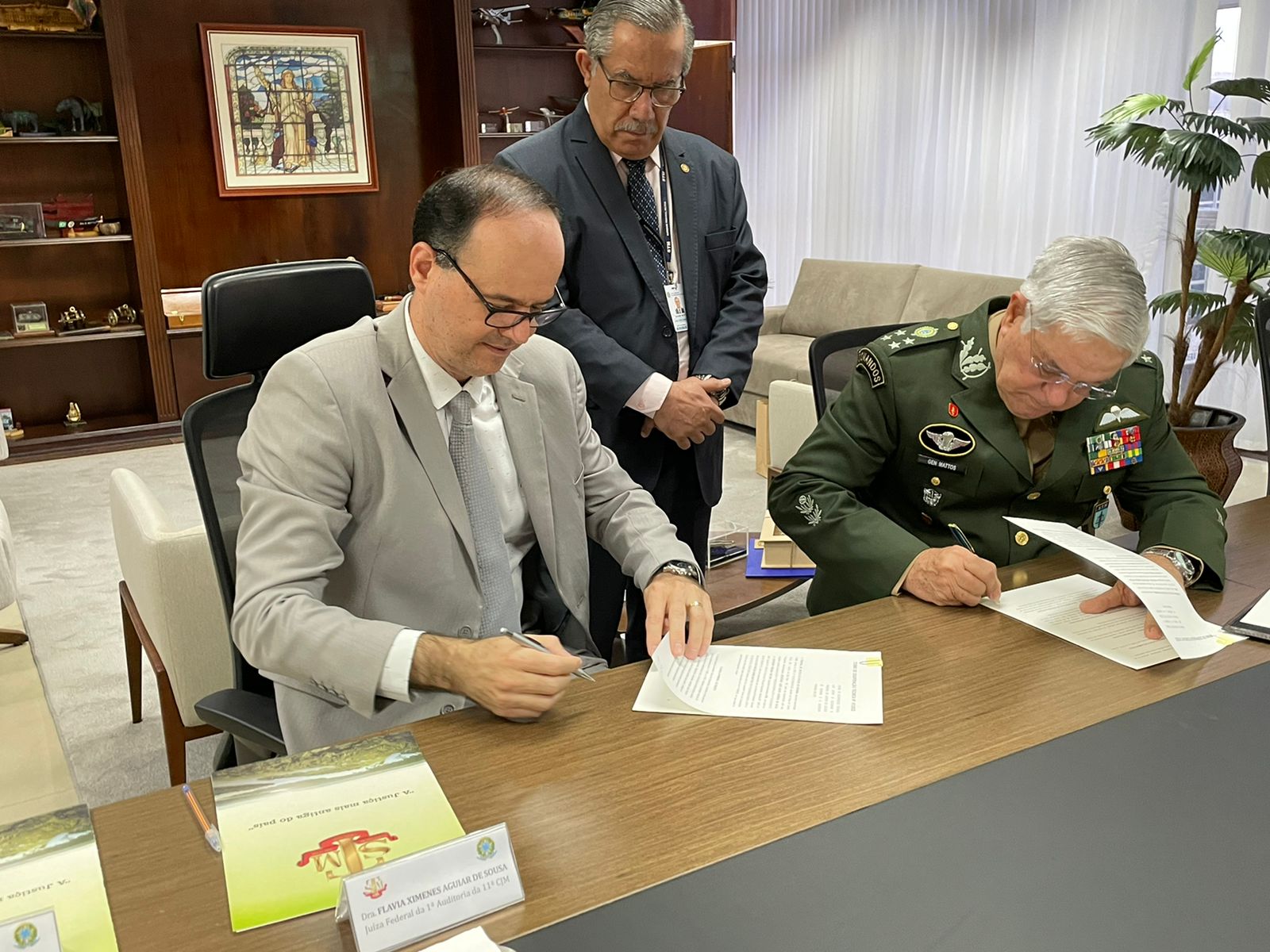 Imagem colorida mostra o presidente do TJRR, sentado, com uma caneta na mão direita e segurando um documento com a mão esquerda, que está em cima de uma mesa. Ao lado está o presidente do STM, também sentado, assinando documento que está em cima de uma mesa. Em pé, atrás dos presidentes, está um homem com as mãos cruzadas. 