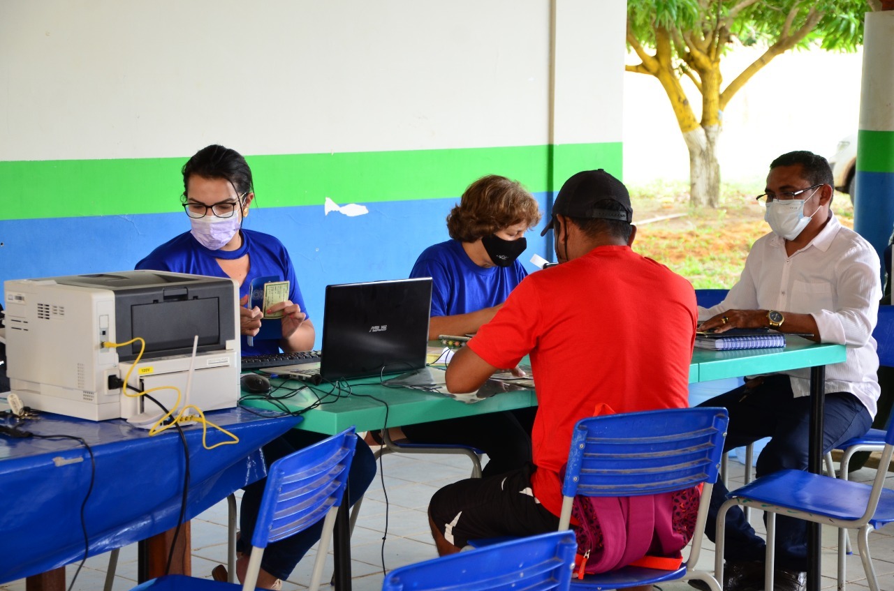 Imagem colorida mostra duas servidoras do TJRR, de camisa azul, sentadas atendendo dois homens, um de camisa vermelha e boné, o outro de camisa branca e calça jeans. Ambos estão sentados em frente a uma mesa com um computador, cabos, cadernos e impressora em cima.