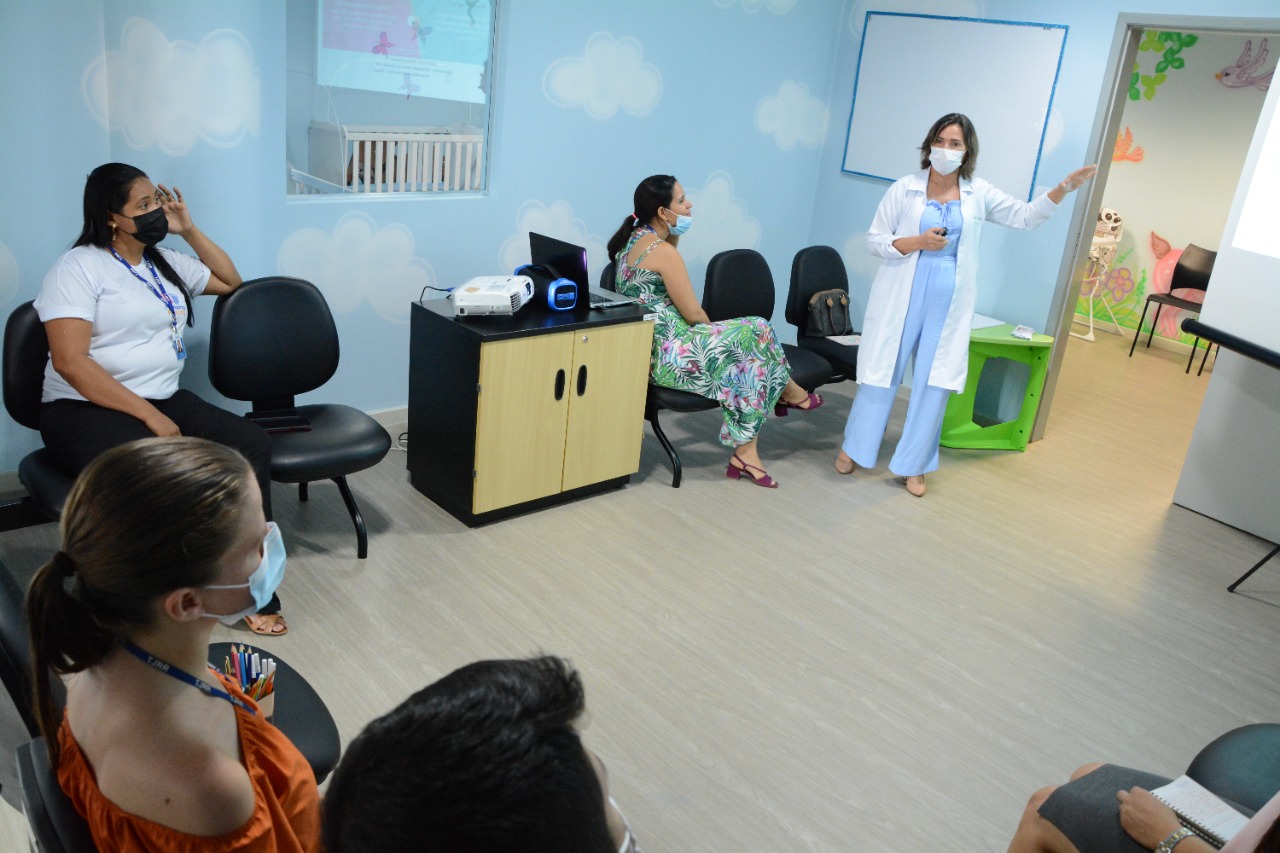  Foto colorida contém pessoas sentadas assistindo palestra na oficina do Programa Primeiros Passos do TJRR, com palestrante em pé apontando para slide projetado na tela. 