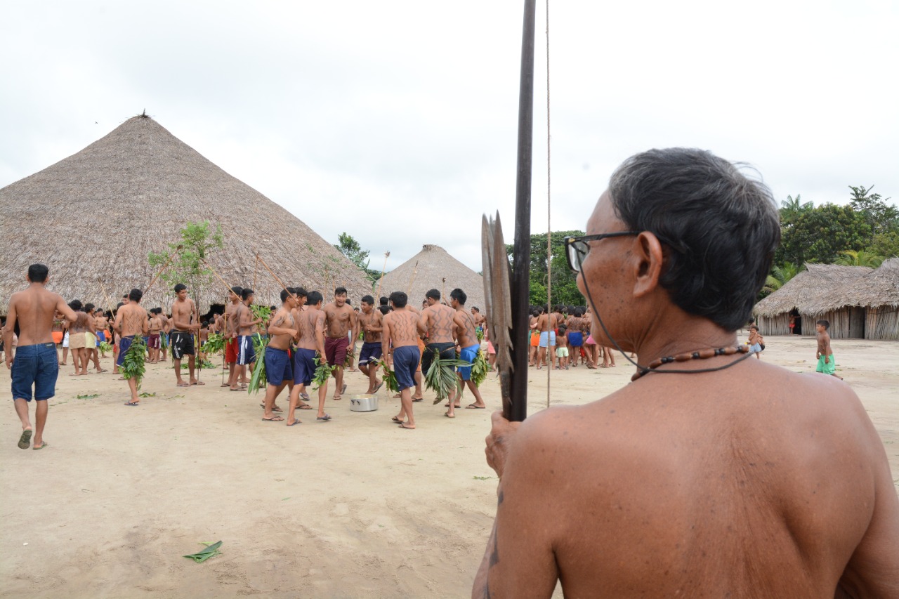 Imagem colorida contém indígena idoso segurando arco e flecha. No fundo da imagem um grupo de homens indígenas dançando durante o ritual de recepção