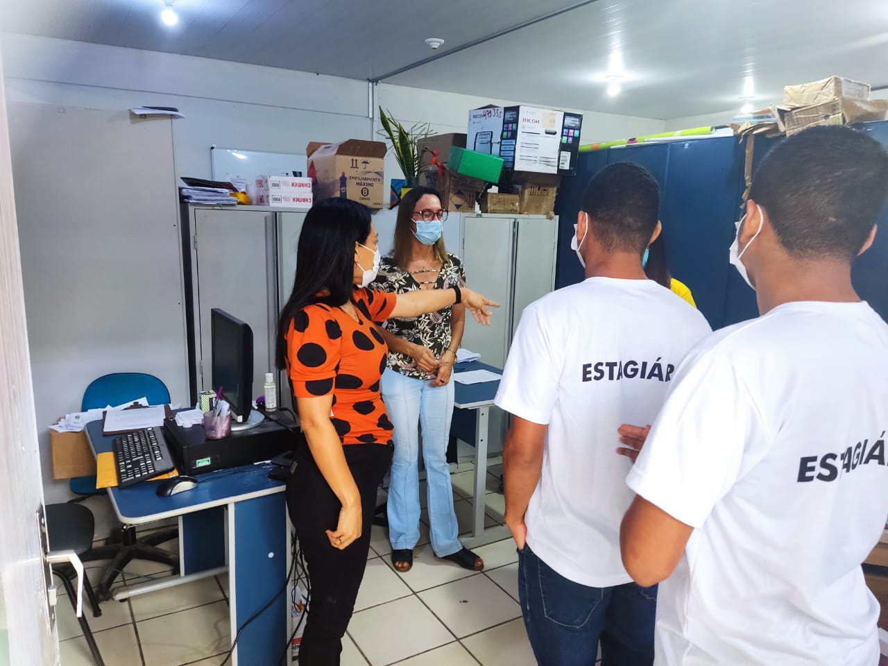 Imagem colorida mostra dois estagiários de camisa branca de costas, duas servidoras da Setrabes, trajando calça jeans e camisa estampada, em pé dentro de uma sala. No local tem mesas, um computador, armários de ferro cinzas e azuis e objetos como papéis e caixas em cima do armário.