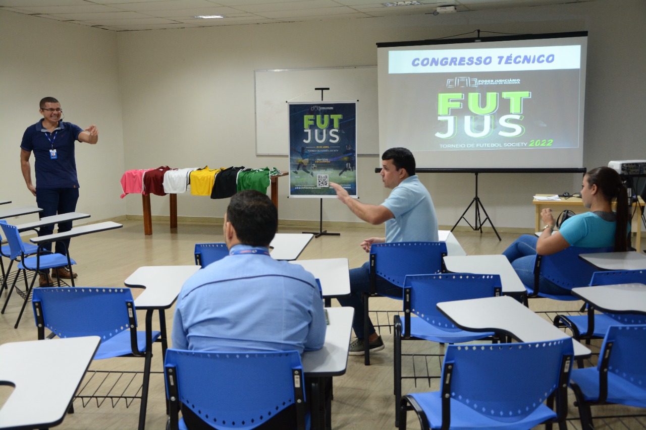 Imagem colorida mostra sala com três pessoas sentadas em cadeiras azuis e o presidente da comissão do evento em pé falando ao microfone. Ao fundo está uma mesa com camisas coloridas em cima, um banner e um projetor com a imagem de divulgação da programação.