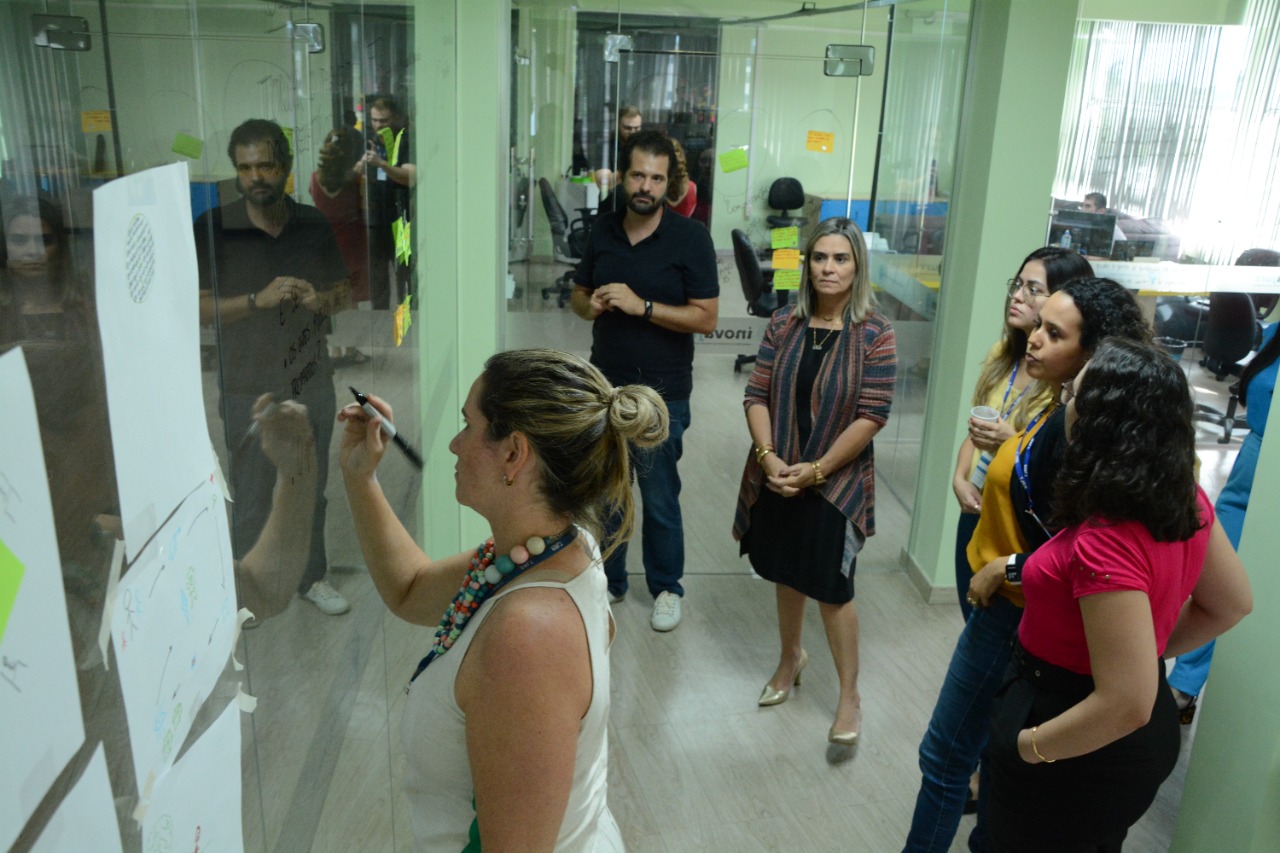 foto colorida mostra uma mulher escrevendo com pincel em quadro de vidro. Outras quatro mulheres e um homem lêem as palavras escritas no quadro. Todos estão em pé.