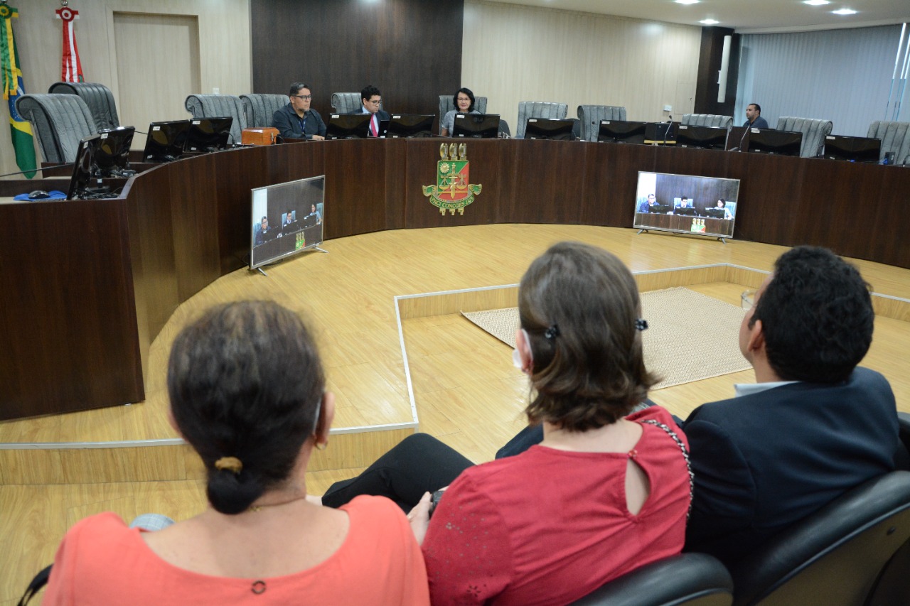 foto colorida mostra três pessoas de costas sentadas na platéia do Tribunal Pleno do Palácio da Justiça. À frente estão sentados a desembargadora Tânia Vasconcelos, o juiz Breno Coutinho e um servidor do TJRR frente a computadores