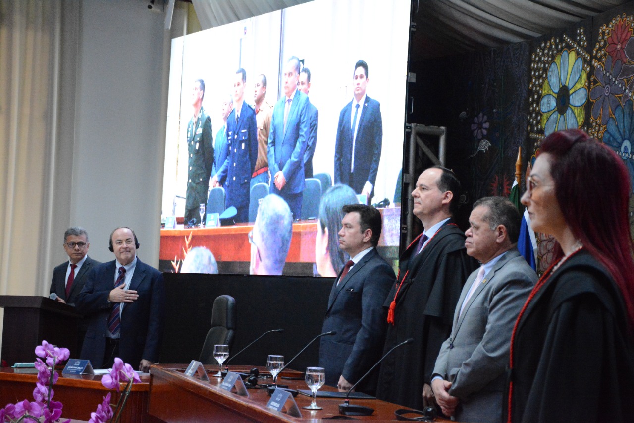 Foto mostra procuradora geral do MPRR, vice-presidente da ALERR, presidente do TJRR, procurador geral do Estado de Roraima, juiz membro da Suprema Corte do Arizona e mestre de cerimônia do evento. Todos em pé durante a execução do hino nacional dos Estados Unidos. 