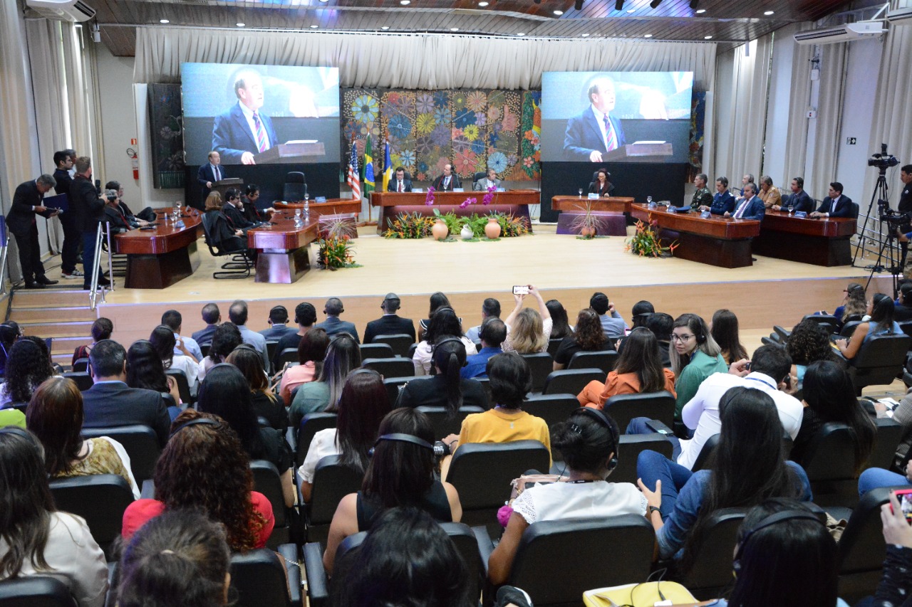 foto colorida mostra pessoas de costas sentadas, utilizando fone de ouvido, frente ao palco do Fórum Cível, onde o palestrante está apresentando de pé. Autoridades do poder público estão no palco sentadas em mesas marrons com flores em frente.