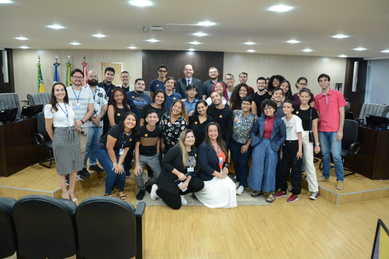 foto colorida mostra 21 estudantes de comunicação social da UFRR posando para foto com o presidente do TJRR, juntamente aos servidores do Tribunal, no Tribunal Pleno. Atrás estão objetos como bandeiras, cadeiras e computadores.