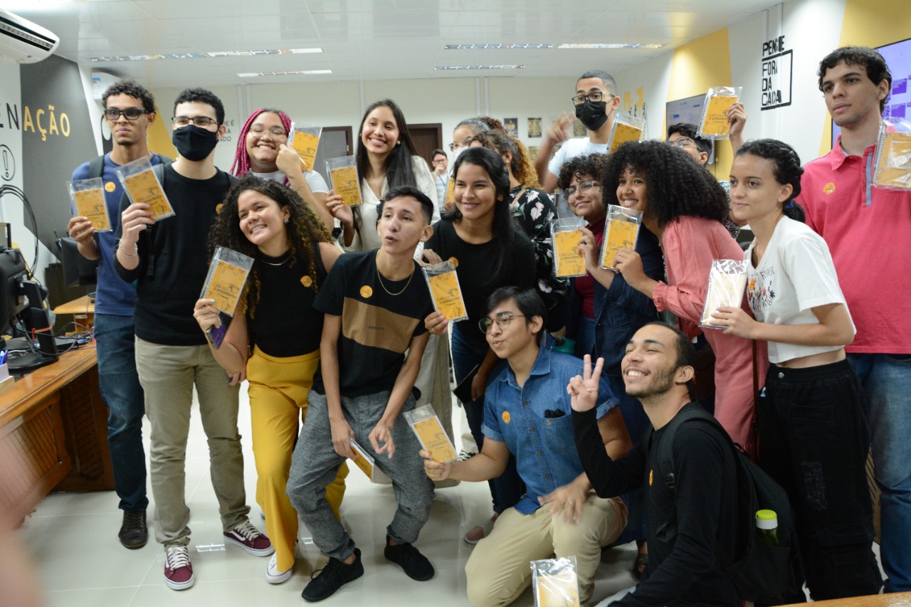 foto colorida mostra 17 estudantes de comunicação social da UFRR segurando um pacote plástico com caderno amarelo e caneta dentro, na sala do NUCRI/TJRR, que possui paredes brancas, amarelas e pretas.