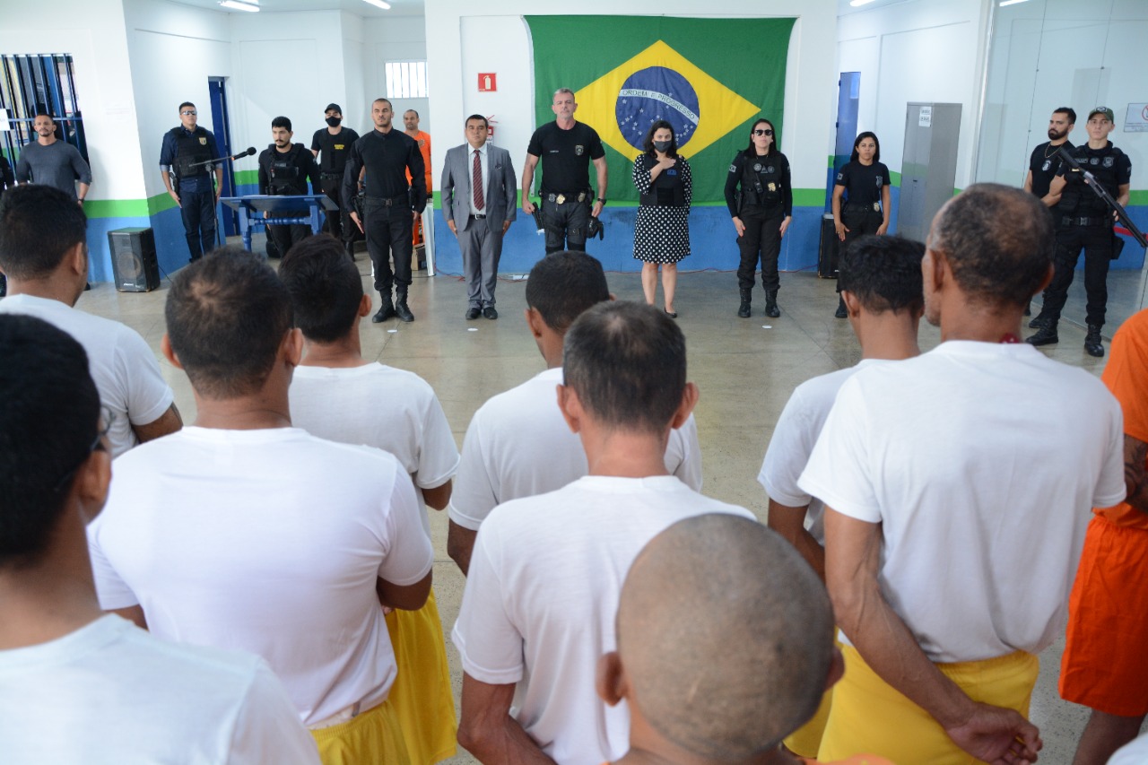 Imagem colorida durante solenidade na cadeia pública masculina, com os detentos de costas pra câmera e logo a frente deles, a  juíza titular da Vara de Execução Penal do Tribunal de Justiça de Roraima (TJRR), Joana Sarmento, com a mão direita no coração, durante hino nacional brasileiro, juntamente com a equipe da unidade prisional.