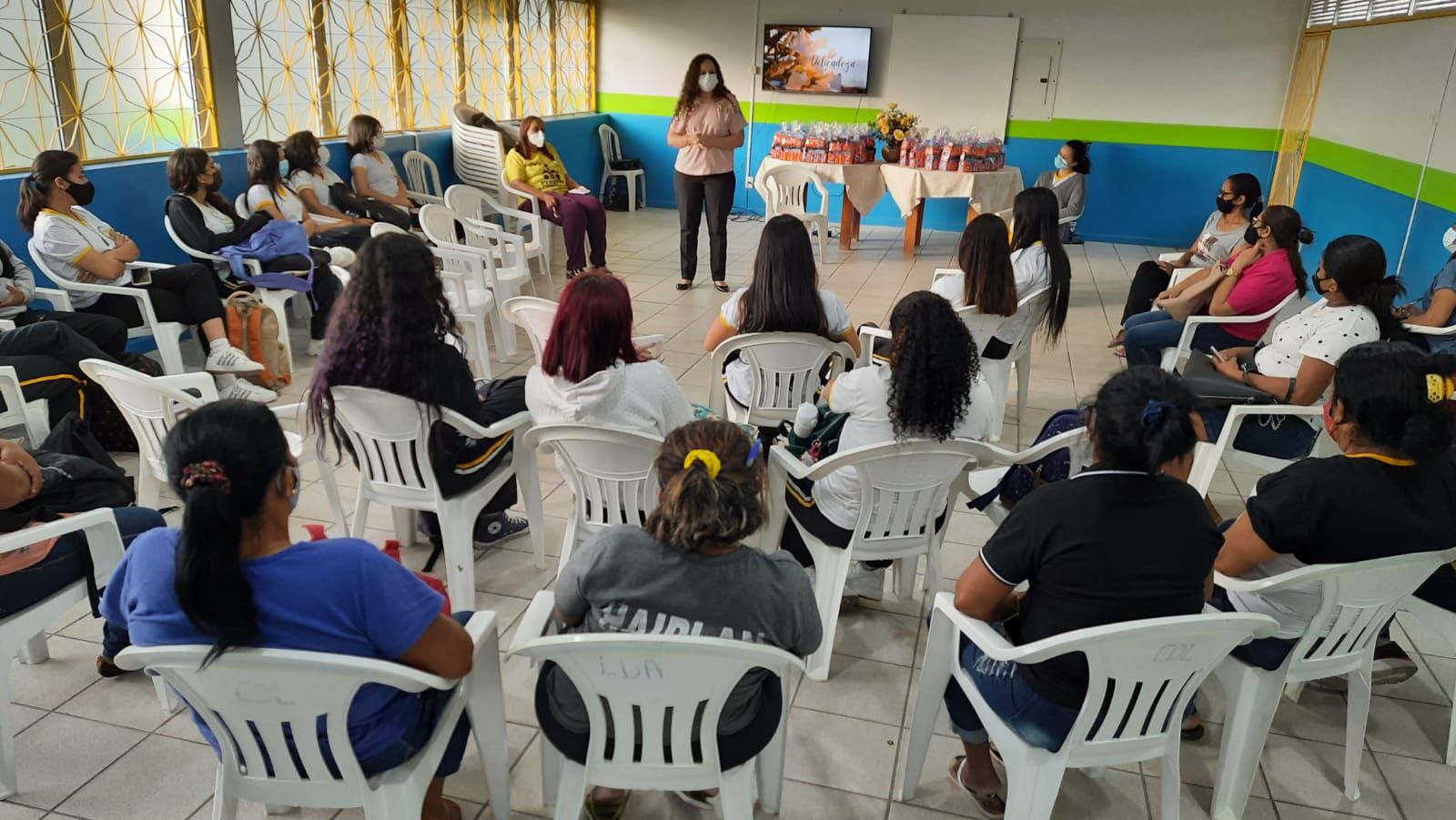 foto colorida mostra cerca de 20 pessoas sentadas em cadeiras brancas assistindo à palestra. A frente está a palestrante ao lado de uma mesa onde está posto um jarro de flores e brindes.