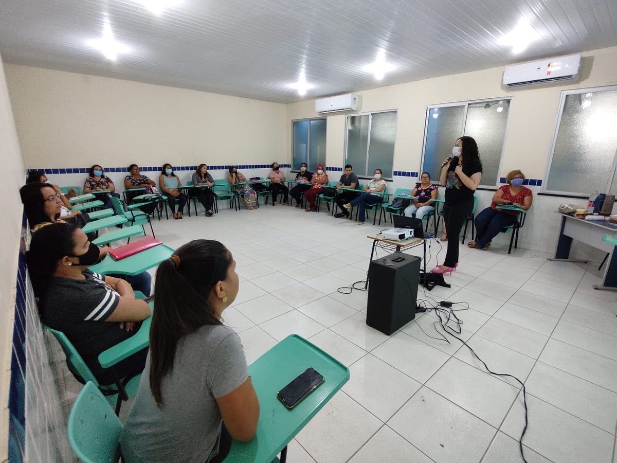 foto colorida mostra 16 pessoas sentadas em cadeiras verdes organizadas em formato “U” em uma sala de aula, assistindo à palestra. A frente está a palestrante falando ao microfone ao lado de uma mesa com notebook e um projetor. Ao lado está uma caixa de som.