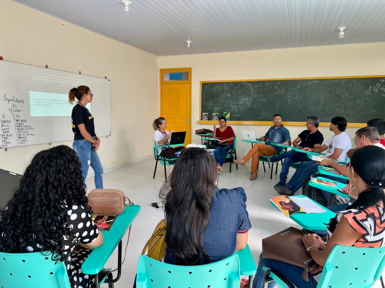 foto colorida mostra servidora do TJRR, Aurilene Mesquita, em pé, trajando calça preta e calça jeans ministrando oficina de capacitação em São Luiz. Na sala de aula estão 10 pessoas sentadas em cadeiras verdes. Objetos como computador, livros e dois quadros compõem a imagem.