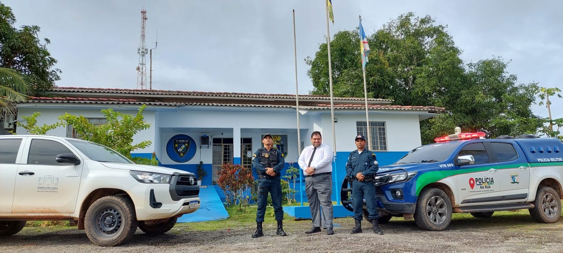 foto colorida mostra dois policiais militares, trajando farda, e o juiz titular da comarca de São Luiz do Anauá, trajando calça jeans, blusa branca e gravata preta, em pé posando para foto. Ao lado direito está uma viatura da PM e ao lado esquerdo um carro oficial do TJRR, ambos estacionados. Todos estão em frente ao prédio da PM em São Luiz 