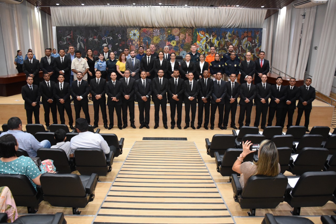 foto colorida mostra 26 pessoas sentadas em poltronas pretas de frente para o palco do auditório no Fórum Cível Advogado Sobral Pinto. No palco estão posicionadas em formato de “U” mesas de madeira envernizada. Nas três principais mesas ao centro estão sentados magistrados e servidores do TJRR e militares do Corpo de Bombeiros e Polícia.