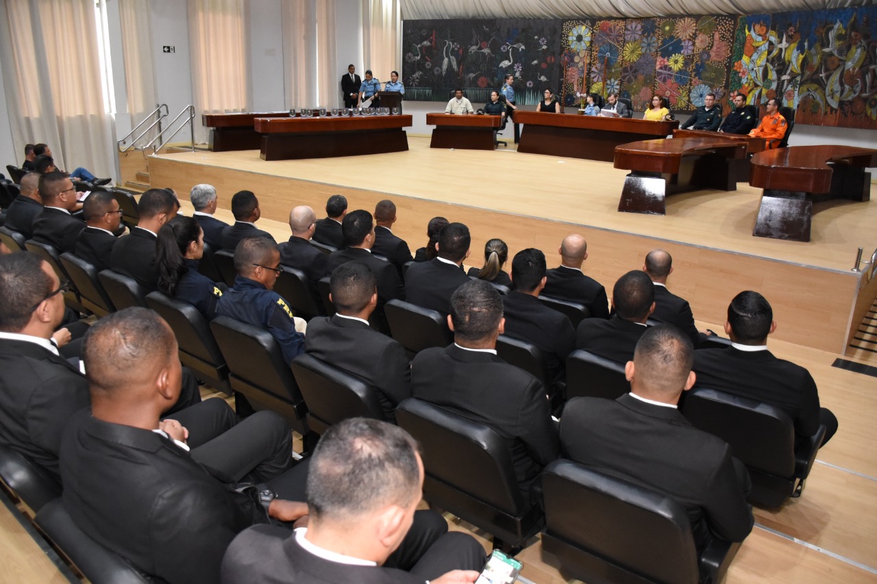 foto colorida quadrada mostra 46 pessoas em pé posando para foto no auditório do Fórum Cível Advogado Sobral Pinto. 19 homens trajando terno, gravata, calça preta e camiseta branca estão em frente ao palco. 27 pessoas, entre homens e mulheres servidores, magistrados e profissionais do Gabinete Militar do TJRR estão em cima do palco. Atrás está uma pintura abstrata colorida grande. À frente das pessoas que posam para a foto estão quatro outras cinco pessoas sentadas em poltronas pretas. 