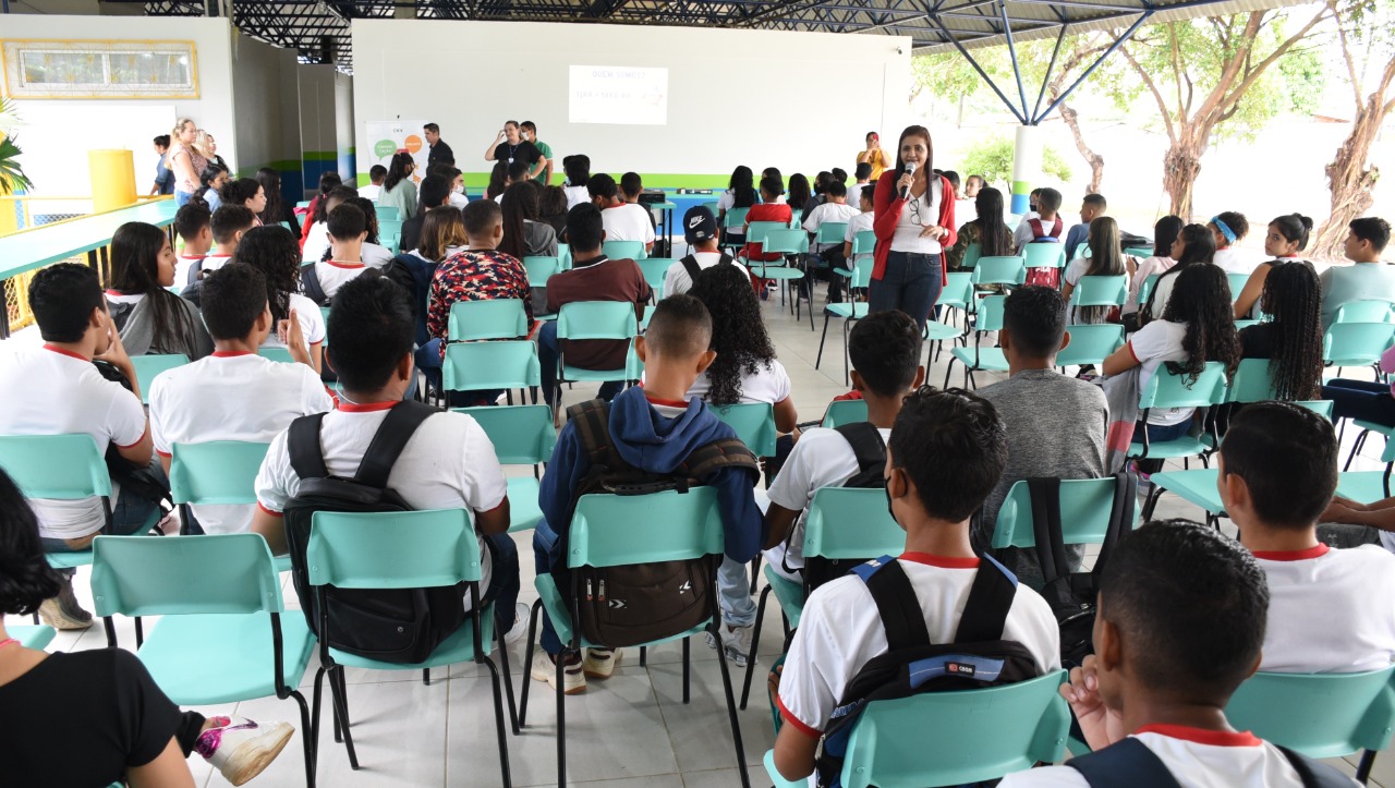 Foto colorida mostra cerca de 50 estudantes sentados em cadeiras verdes na área de convivência da Escola Estadual Olavo Brasil. Todos estão de costas para a foto. Entre os alunos está a palestrante Renatta Alves em pé, falando ao microfone, de frente para os alunos. A palestrante traja calça jeans, blusa branca e casaco rosa. Os alunos vestem farda branca com gola vermelha