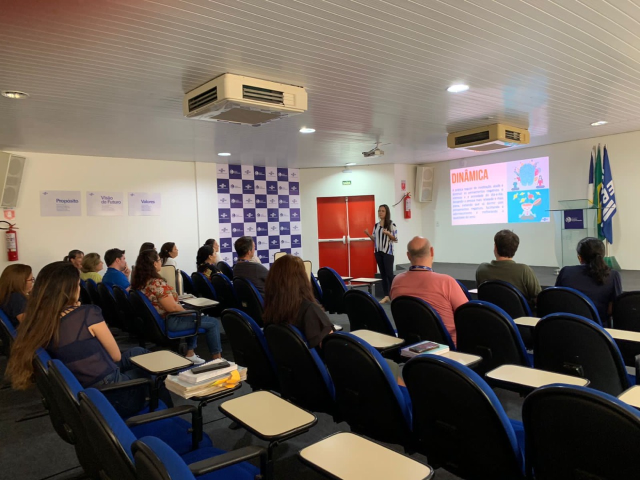 A imagem colorida quadrada mostra cerca de 15 colaboradores do Sebrae Roraima sentados em poltronas azuis de auditório. Frente ao palco está a palestrante em pé, trajando calça preta e camisa listrada nas cores preta e branca. Atrás, na parede, está projetado um slide. Ao lado estão três bandeiras e um púlpito.