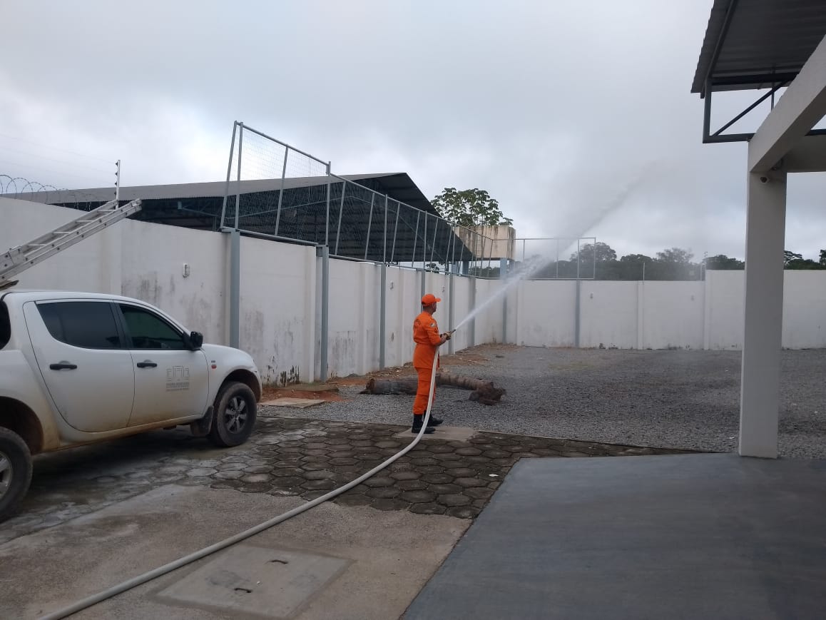 Imagem colorida contém integrante da equipe do Corpo de Bombeiros do Gabinete Militar (Gabmil) manobrando a mangueira de incêndio em área aberta, durante ação de prevenção. Ao lado dele contém carro do TJRR. 