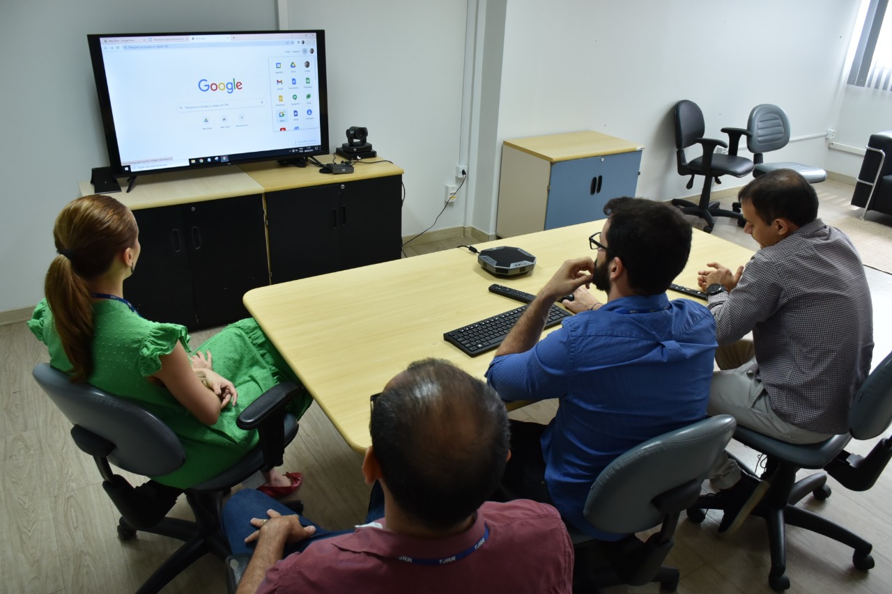  Imagem colorida contém quatro servidores da Secretaria de Tecnologia da Informação do Tribunal de Justiça de Roraima (STI/TJRR)  sentados em frente a uma smart tv durante demonstração da aquisição de novas ferramentas do Google (empresa multinacional de serviços online e software). 