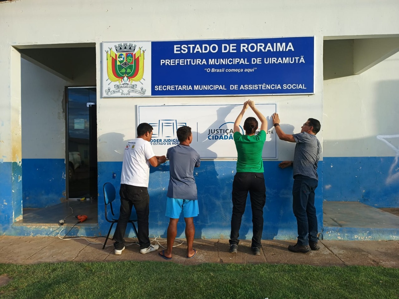 imagem contém quatro pessoas arrumando a placa de identificação do Polo do Justiça Cidadã, na prefeitura municipal de Uiramutã. 