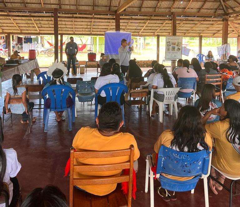 foto colorida mostra cerca de 20 mulheres indígenas da comunidade da Barata assistindo palestra ministrada pelo juiz Parima Dias Veras, que discursa em pé com um microfone na mão esquerda. Ambas estão embaixo de um malocão.Todos estão debaixo de um malocão.