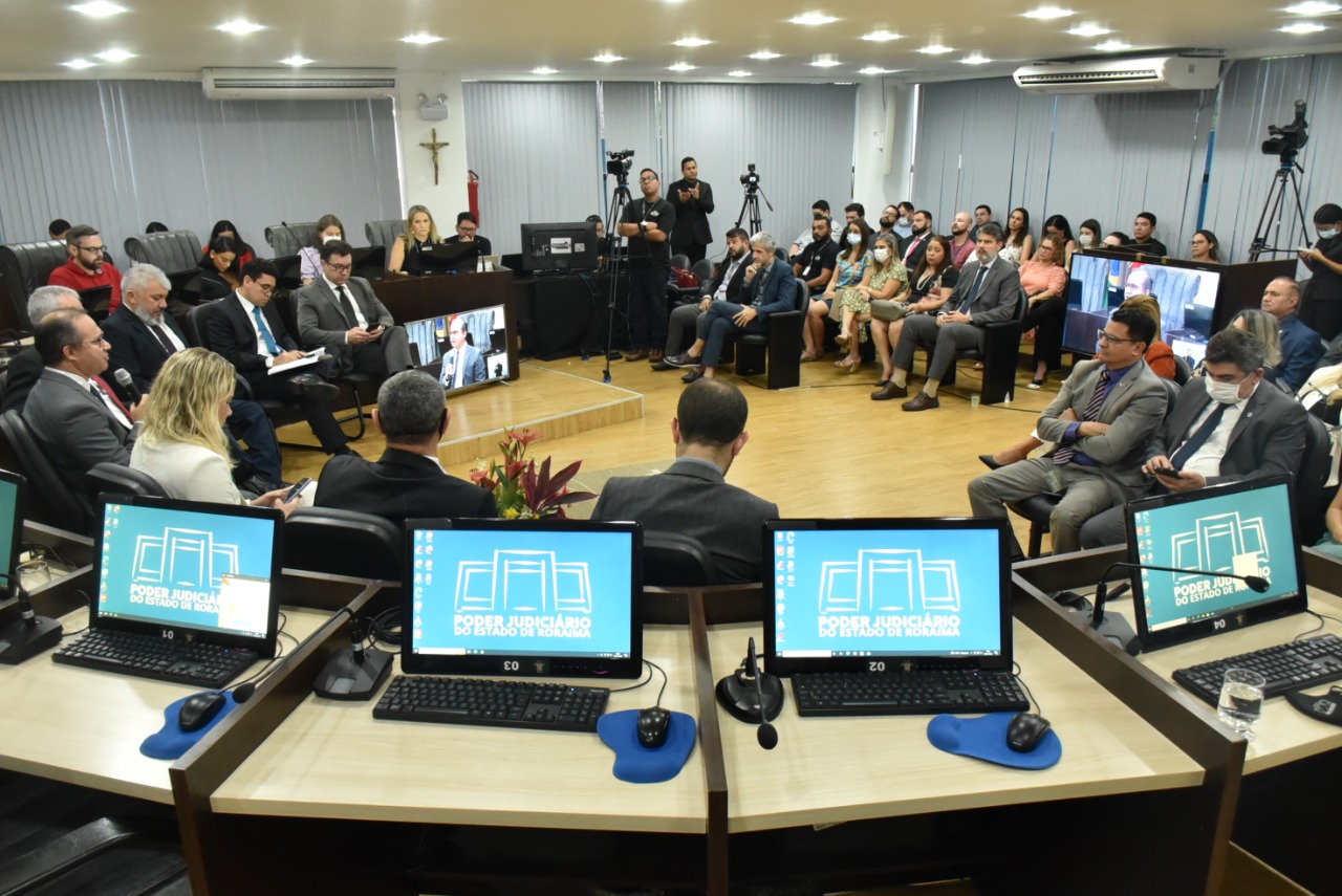 Foto colorida mostra sala de audiências do Tribunal Pleno durante audiência pública. Em primeiro plano, aparecem telas de quatro computadores que ficam nas tribunas. À frente estão sentados magistrados do TJRR, autoridades da OAB-RR, Defensoria Pública e demais autoridades do poder público roraimense. Adiante está a plateia, sentada de frente para as autoridades. Um intérprete de libras aparece em pé, frente a uma câmera ao lado direito do auditório. Objetos como câmeras, televisores e centrais de ar também aparecem na imagem. 