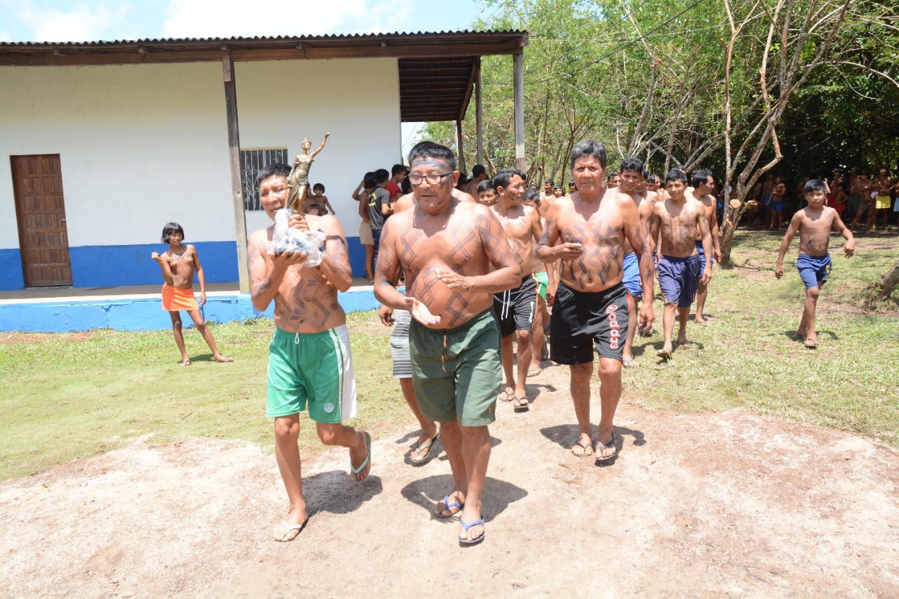 Imagem colorida contém o povo Waimiri Atroari durante ritual de recepção aos integrantes do TJRR. Na imagem um dos homens indígenas carrega a estátua da justiça. 