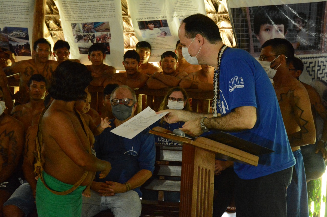 Imagem colorida contém o presidente do TJRR, desembargador Cristóvão Súter, entregando nas mãos de uma mulher indígena documentação, durante cerimônia dentro da Maloca onde indígenas se reúnem na comunidade. 