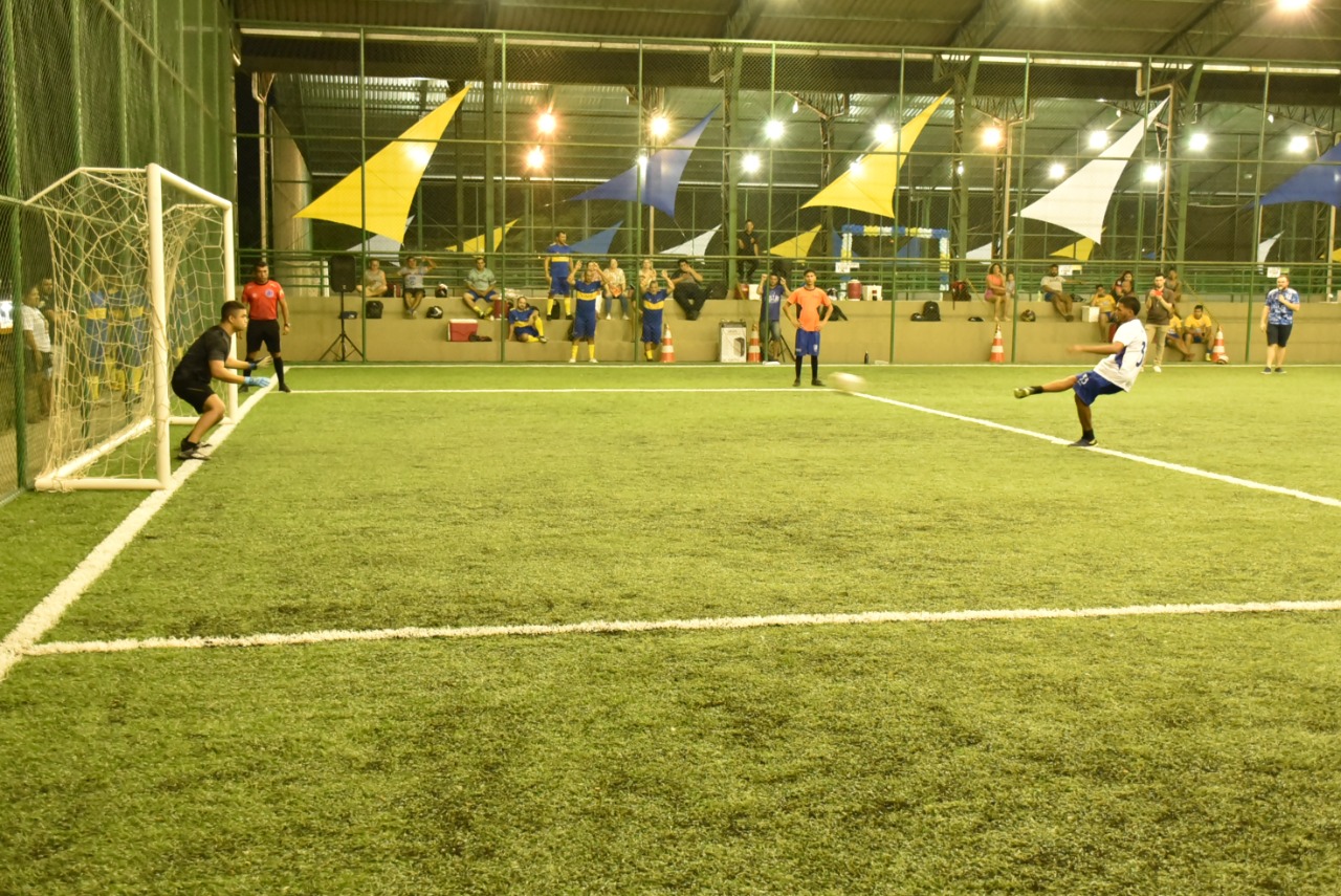 Imagem colorida contém jogador de futsal chutando a bola na direção da rede e o goleiro em posição de defesa. 