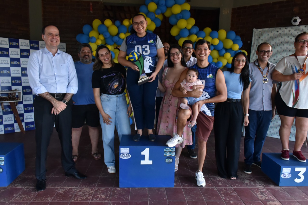 Imagem colorida contém  participantes dos torneios Futjus e voleijus posando para fotografia no pódio de primeiro lugar ao lado do presidente do TJRR, desembargador Cristóvão Súter. Na imagem, um dos participantes segura uma criança de colo nos braços. 