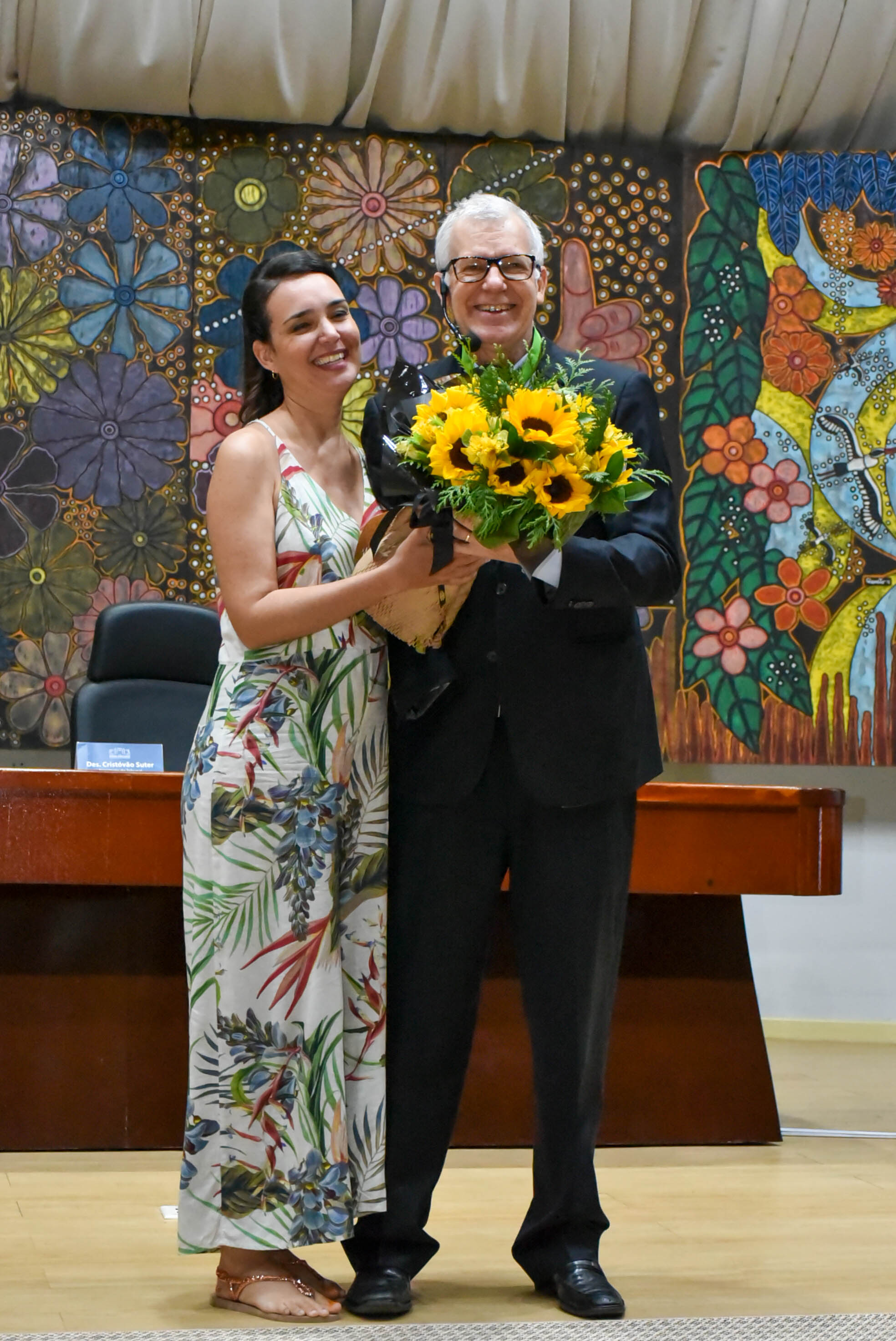  Imagem colorida contém o ministro do STJ, recebendo das mãos da servidora do TJRR, Rudianna Zeidler,  um buquê de flores. 