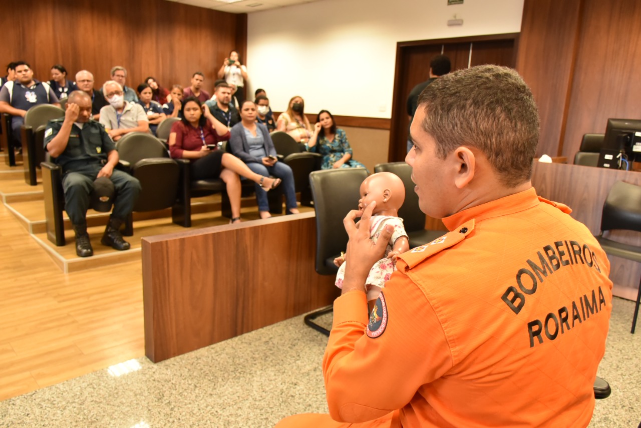 Imagem colorida do Primeiro Tenente Fernandes, segurando boneca para simular crianças, durante palestra no Fórum Criminal sobre primeiros socorros. 