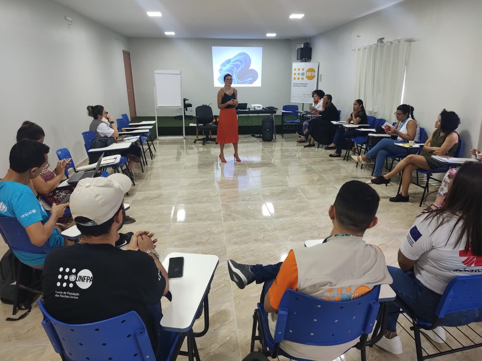 Imagem colorida contém pessoas reunidas em uma roda de conversa. No meio da roda, encontra-se a palestrante. 