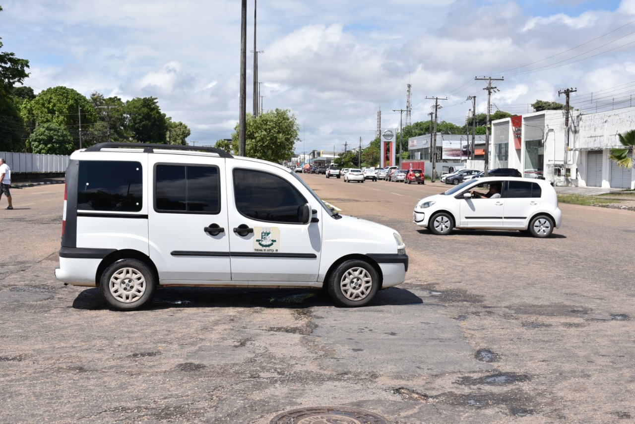 Imagem colorida mostra um carro modelo Fiat uno com o selo do tribunal de justiça estacionado próximo a uma via com carros em movimento