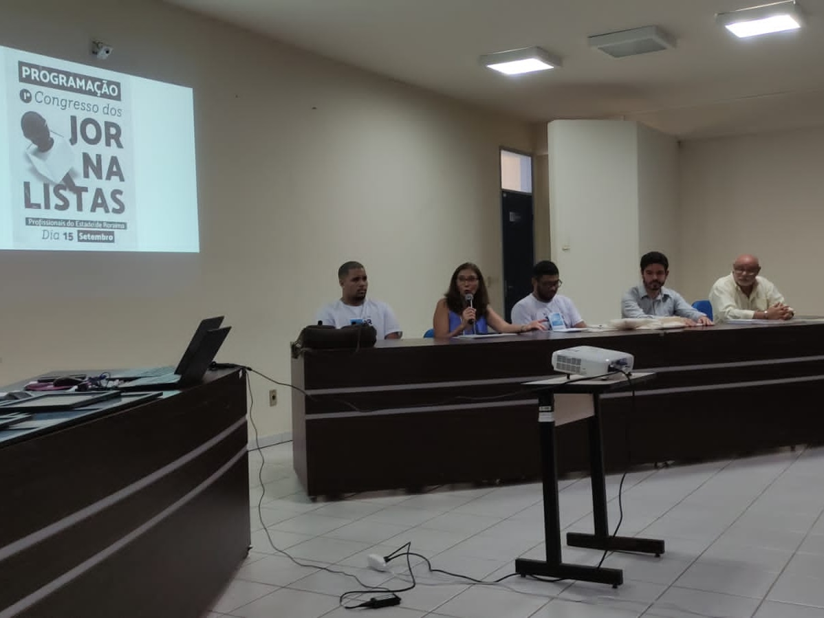 Imagem colorida mostra uma bancada com 4 pessoas sentados, sendo que uma delas está com o microfone na mão, à frente tem um projetor e na lateral esquerda há uma segunda bancada com papéis, quadros e notebooks