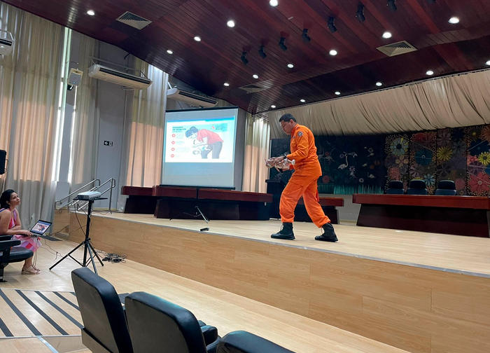 Imagem colorida do facilitador 1º Tenente Fernandes  usando um uniforme laranja em um auditório, segurando uma boneca . 