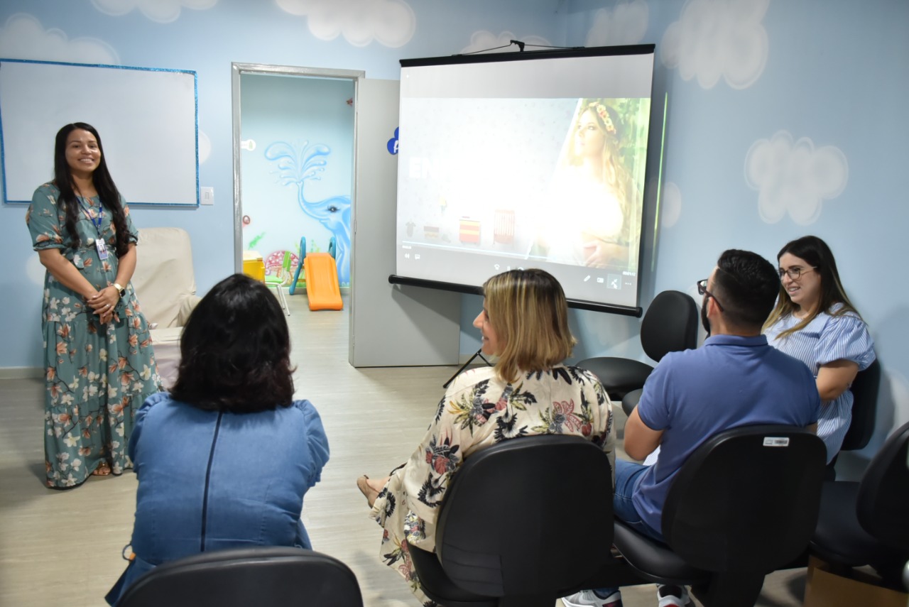 Imagem colorida contém cinco pessoas, sendo 4 delas sentadas de frente para a palestrante que se encontra em pé. Atrás da palestrante encontra-se tela de apresentação para power point. 