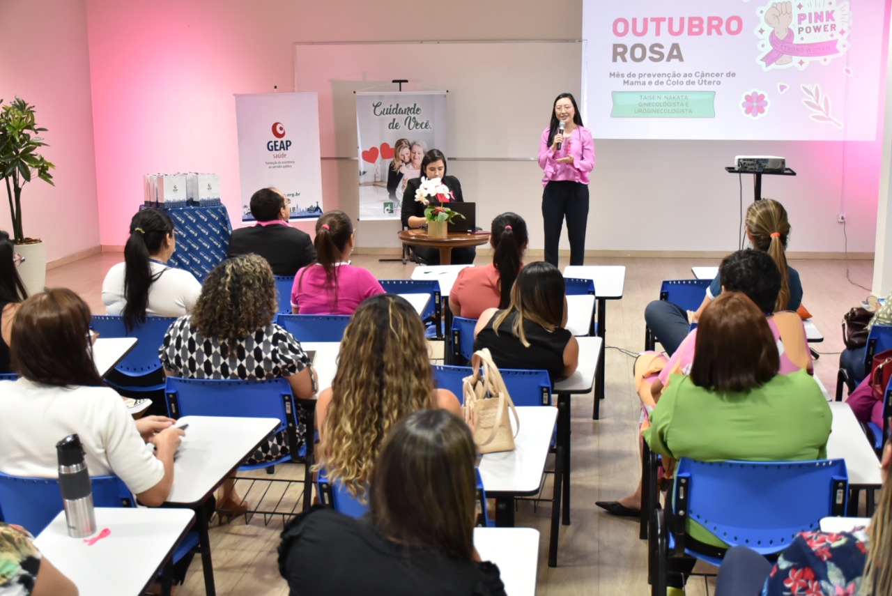 Imagem colorida contém a ação do Outubro Rosa com a palestrante em pé de frente para servidoras do TJRR na Escola do Poder Judiciário de Roraima. Ao fundo, banner da GEAP e sala iluminada com luz rosa. 