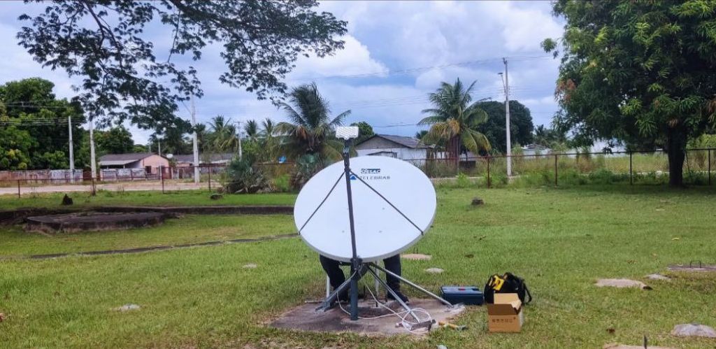 Imagem colorida em formato retangular mostra uma antena redonda sendo instalada em um campo gramado, ao fundo há árvores e algumas residências.