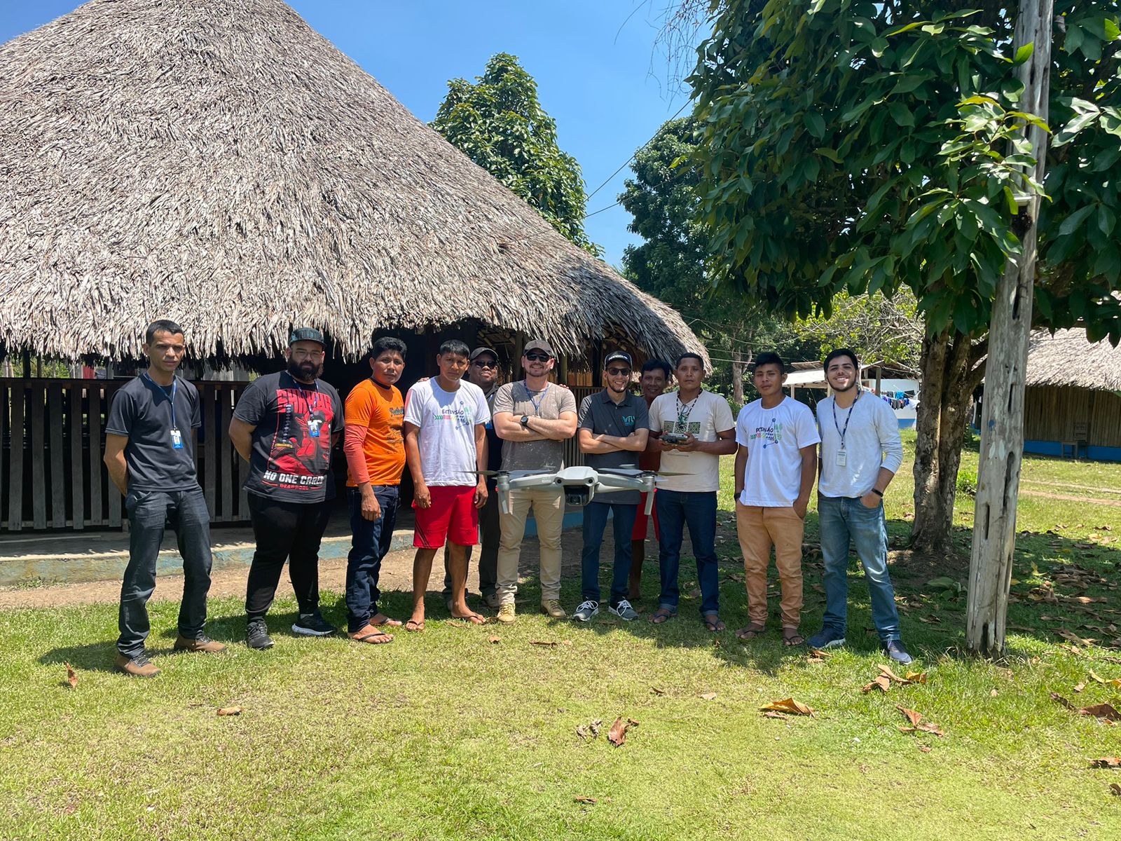 Imagem colorida mostra 11 homens e um drone pairando no ar, entre eles há  servidores do TJRR e indígenas da comunidade Waimiri-Atroari, os mesmos estão em pé posando para a foto em frente a um malocão.