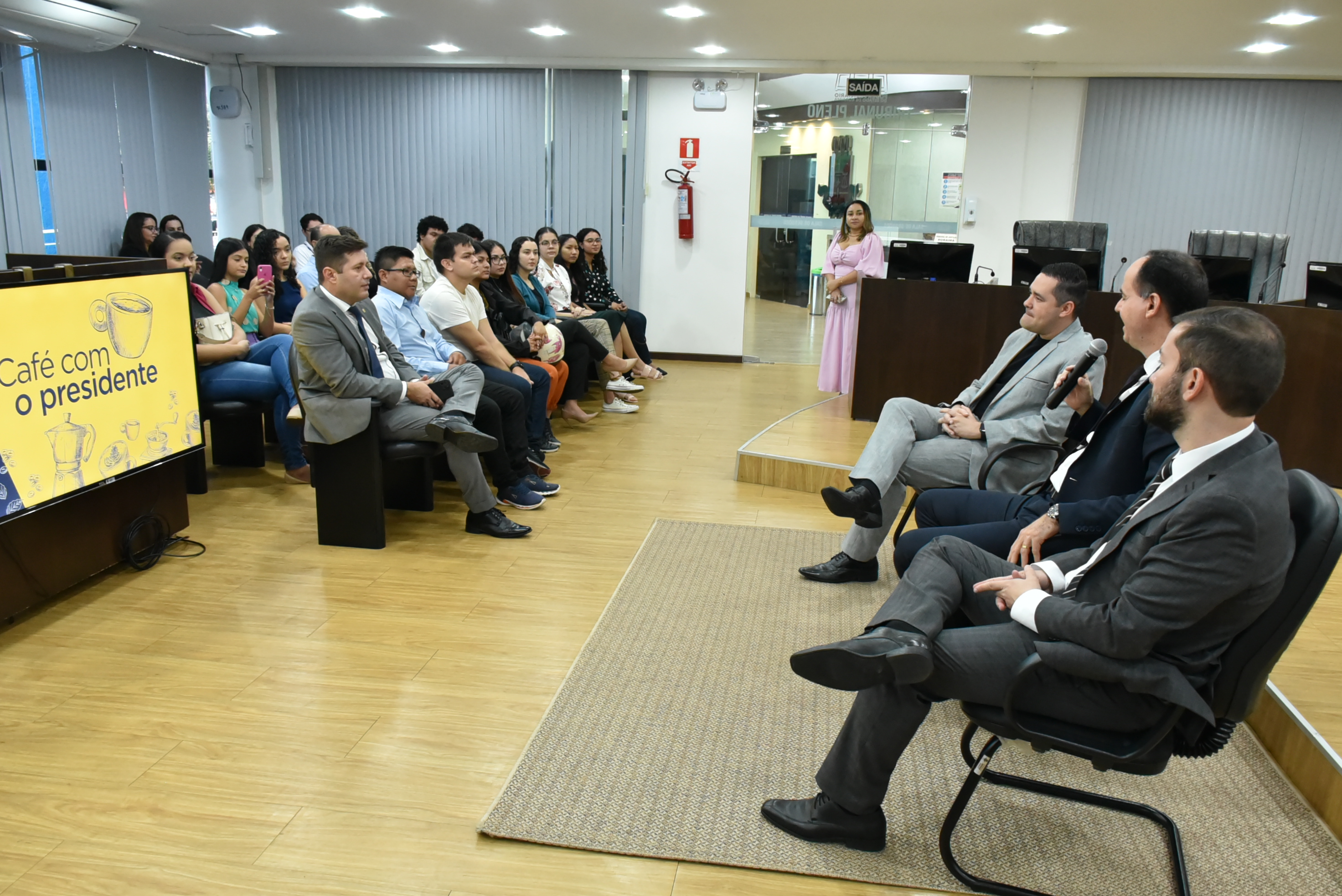 Imagem colorida em formato retangular mostra um grupo de alunos, alunas e  professores da UFRR sentados no auditório da sala do tribunal pleno, a frente também sentado e conversando com os visitantes está o presidente do TJRR, desembargador Cristóvão Suter, acompanhado do secretário geral de justiça, Felipe Queiroz, e do juiz auxiliar da presidência, Bruno Costa. No lado esquerdo da imagem tem um cartaz amarelo com a logomarca do evento “Café com o Presidente”.