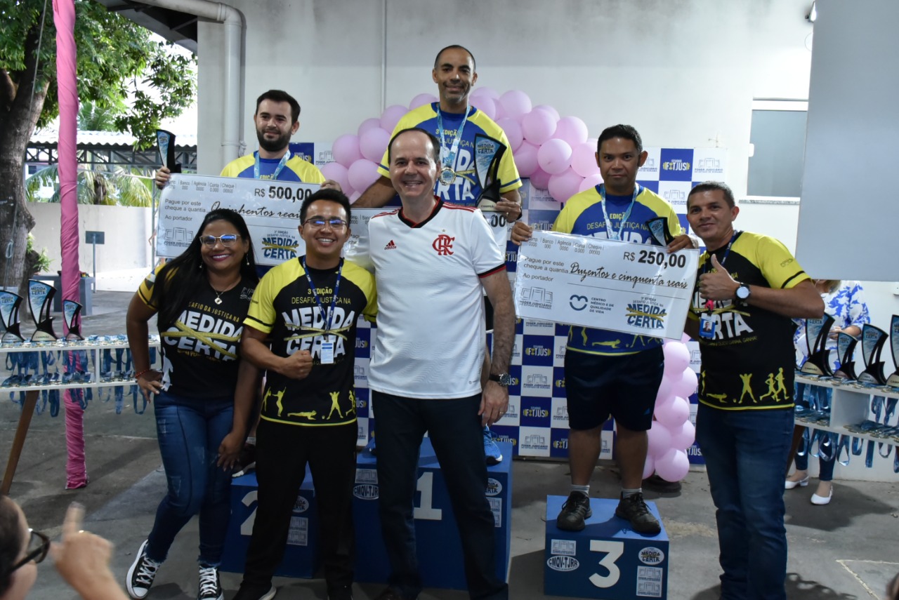  imagem colorida mostra o presidente do TJRR, desembargador Cristóvão Suter, acompanhado dos três vencedores da categoria masculina de perda de percentual de gordura corporal  e dos educadores físicos do TJ. Os vencedores encontram-se em cima do podium com suas medalhas e com um cheque representando o valor de sua premiação. 