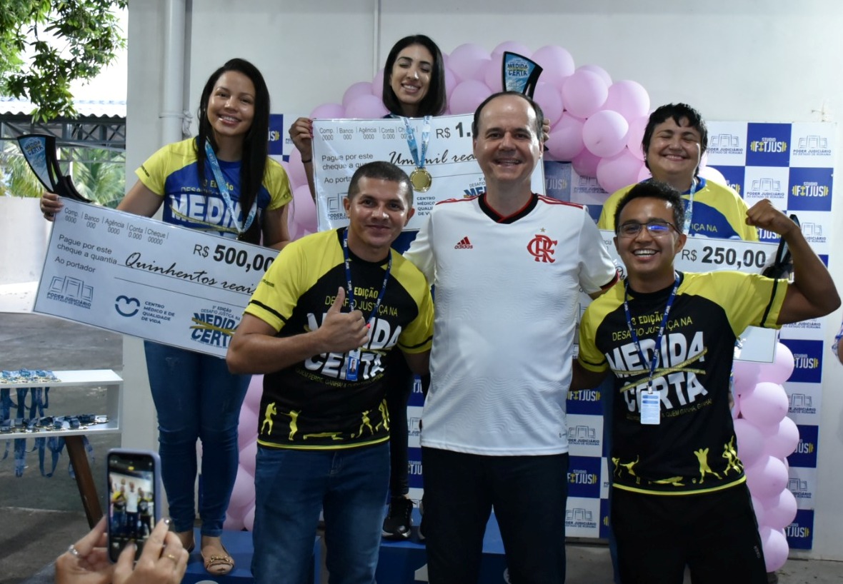 imagem colorida mostra o presidente do TJRR, desembargador Cristóvão Suter, acompanhado das três vencedoras da categoria feminina  de perda de percentual de gordura corporal e dos educadores físicos do TJ. As vencedoras encontram-se em cima do podium com suas medalhas e com um cheque representando o valor de sua premiação. 