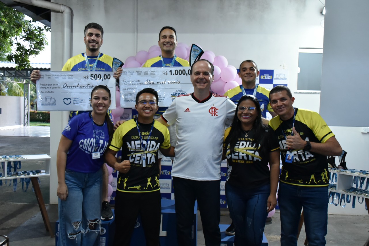 imagem colorida mostra o presidente do TJRR, desembargador Cristóvão Suter, acompanhado dos três vencedores da categoria masculina de ganho percentual de massa muscular esquelética  e dos educadores físicos do TJ. Os vencedores encontram-se em cima do podium com suas medalhas e com um cheque representando o valor de sua premiação. 