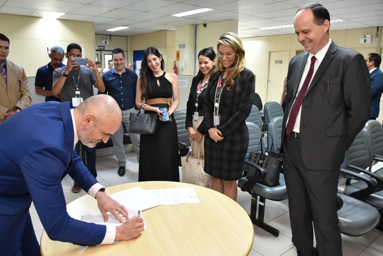 imagem colorida em formato retangular mostra o juiz coordenador dos Juizados Especiais, Air Marin assinando o termo de cooperação em uma mesa redonda, á sua frente está o presidente do TJRR, desembargador Cristóvão Suter e a diretora de relações institucionais e gerente do departamento jurídico da Roraima Energia, Sarassele Freire, juntamente com um grupo de pessoas prestigiando o feito.