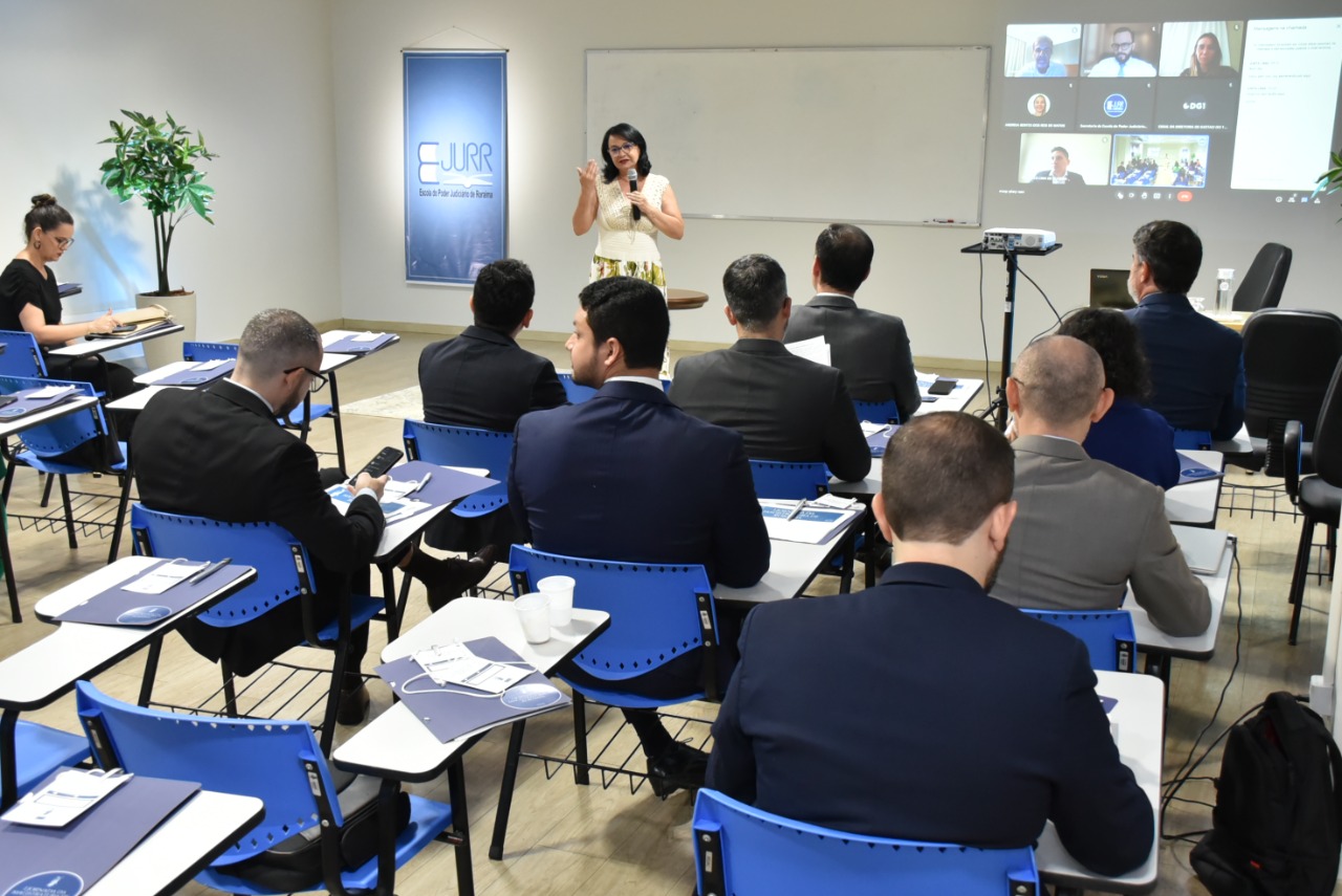  Imagem colorida mostra os magistrados e magistradas sentados em carteiras de tons azul e branco dentro de uma sala com um quadro branco ao lado do quadro há uma projeção de uma sala virtual com oito participantes, na  frente do quadro está a mediadora do evento em pé  com o microfone na mão. 