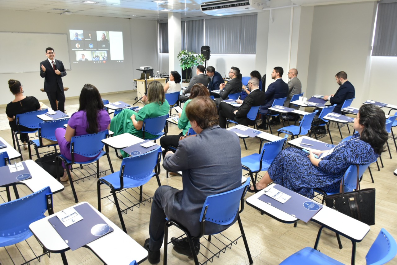 Imagem colorida mostra os magistrados e magistradas sentados em carteiras de tons azul e branco dentro de uma sala com um quadro branco ao lado do quadro há uma projeção de uma sala virtual com oito participantes, na  frente do quadro está o mediador do evento em pé com o microfone na mão. 