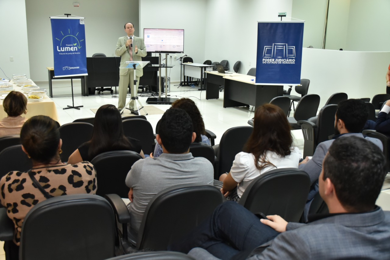 Imagem colorida mostra  o presidente TJRR, desembargador Cristóvão Suter, discursando em um auditório para um grupo de homens e mulheres, a foto mostra também, dois banners azuis, um com a logo do TJRR e outra com a logo do projeto lumen, ambas estão posicionadas nas laterais à frente da plateia.