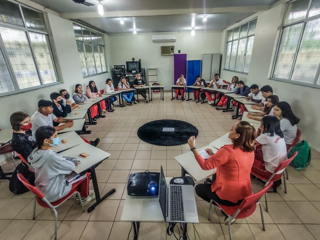  Imagem colorida mostra alunos, alunas,  sentados em  uma sala de aula com carteiras escolares formando um círculo, a facilitadora está sentada entre os estudantes vestindo um blazer no tom laranja, ao seu lado encontra-se uma mesa com um projetor e um notebook sobre ela.