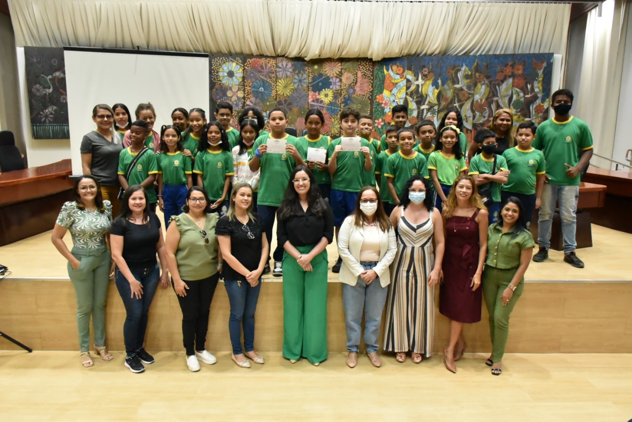  Imagem colorida em formato retangular mostra a coordenadora da Cevid , juíza Suelen Alves e a secretária Adjunta Pedagógica, Meiry Jane Silva posando em pé para foto ao lado de alunos, alunas e professoras da Escola Municipal Ana Sandra Nascimento.